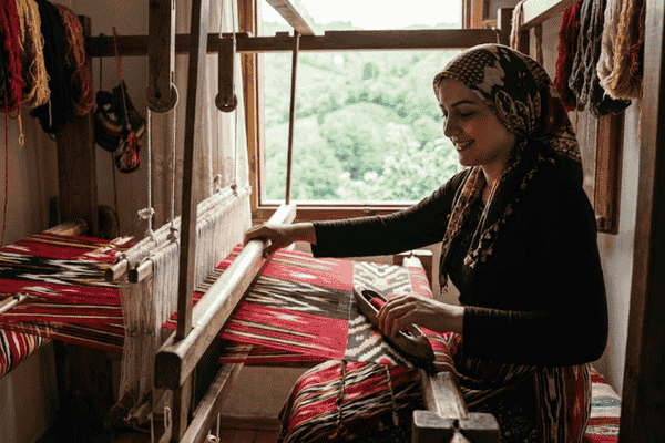 Karadeniz manzarasında, ahşap bir el dokuma tezgahında kırmızı, siyah ve sarı renkli ipliklerle İkat benzeri geleneksel yöntemle Keşan bezi dokuyan, yöresel kıyafetli ve başörtülü gülümseyen bir kadın.