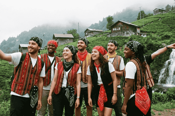 Karadeniz'in sisli ve yeşil yayla manzarasında, arkalarında şelale, ahşap evler ve çay tarlaları varken, üzerlerinde geleneksel kırmızı, siyah ve sarı çizgili Keşan bezinden yapılmış yelekler ve bandanalarla neşeyle grup selfisi çeken yedi kişilik genç gezgin grubu.